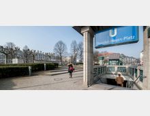 U-Bahn-Eingang zum U-Bahnhof Theodor-Heuss-Platz in Berlin, mit blauem Schild und Treppen, die in den Untergrund führen. Im Hintergrund sind beschnittene Bäume, Hecken, Bürgersteige und Wohngebäude zu sehen.