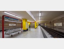 U-Bahn-Station "Jakob-Kaiser-Platz" in Berlin mit gelben Säulen, moderner Sitzbank und einem Hinweisschild der Linie U7. Rechts befindet sich das Gleis mit einer Bahnsteigkante, links führt eine Treppe zur nächsten Ebene.