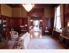 Eleganter Innenraum eines historischen Gebäudes mit Holzwänden, einem dekorativen Kronleuchter und großen Fenstern mit Vorhängen. Im Vordergrund befindet sich eine gemütliche Sitzgruppe mit Sofa und Sesseln, während ein Flur in einen weiteren Raum mit gedeckten Tischen führt.