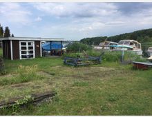 Ein grasbewachsener Bereich am Ufer eines Sees mit einem kleinen braunen Holzschuppen mit weißen Türen links. Im Hintergrund sind mehrere kleine Boote an einem Steg festgemacht, umgeben von Schilf und Bäumen.