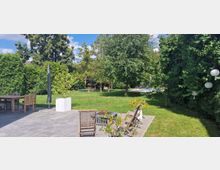 Gepflegter Garten mit einer Terrasse aus Steinplatten, darauf stehen hölzerne Gartenmöbel und ein weißer Blumenkübel. Im Hintergrund befinden sich eine große Rasenfläche, dichte Bäume und Büsche sowie ein Poolbereich.