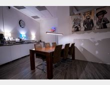 Ein moderner Besprechungsraum mit einem Holztisch und sechs gepolsterten Stühlen, einer Küchenzeile mit Kaffeemaschine und Geschirr im Hintergrund sowie einer Neonleuchte über dem Tisch. An der Wand hängen drei Bilder mit Tierfiguren in Anzügen, und der Boden ist mit dunklem Laminat ausgelegt.