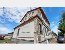 Ehemaliges Gebäude mit der Aufschrift "Lehrlingswohnheim" in heller, verwitterter Fassade und rotem Dach. Im Vordergrund ein Bauzaun, im Hintergrund ein Gehweg mit Autos und Bäumen.