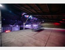 Industriehalle mit Stahlträgerdach und offenem Betonboden, ergänzt durch eine lange Metallbank. Die Wände bestehen aus unverputzten Ziegeln, und bunte Lichter sowie Gerüste im Hintergrund verleihen dem Raum ein modernes, eventorientiertes Ambiente.