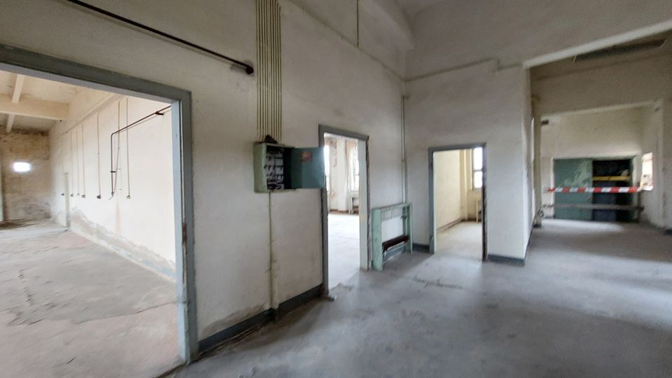 Ein leerer Raum in einem industriellen oder verfallenen Gebäude mit weiß gestrichenen Wänden, sichtbaren Kabeln und offenen Türen, die in angrenzende Räume führen. Im Vordergrund ist ein alter Sicherungskasten zu sehen, und einige Bereiche wirken durch Absperrband abgetrennt.