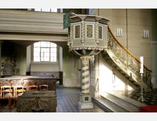 Innenraum einer Kirche mit einer reich verzierten Kanzel auf einer gedrehten Säule, einer geschwungenen Treppe mit dekorativem Geländer und einem Taufbecken im Vordergrund. Im Hintergrund befindet sich ein großes Fenster und hölzerne Kirchenbänke.