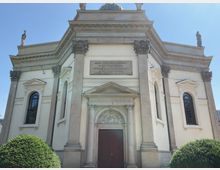 Historische Kirche mit klassizistischer Fassade, verziert mit Säulen, Bögen und Reliefs. Über dem Eingang ist eine Inschrift, und die Fassade wird von Statuen auf den Dachkanten gekrönt; zwei Büsche flankieren den Eingangsbereich.