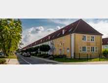 Reihenhäuser mit gelbem Putz und roten Satteldächern entlang einer ruhigen Straße, flankiert von Bäumen und geparkten Autos; ein Gehweg und eine begrünte Vorgartenfläche sind sichtbar. Im Hintergrund sind weitere Gebäude und ein leicht bewölkter Himmel erkennbar.