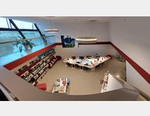 Innenraum einer modernen Bibliothek mit hohen Decken, großen schrägen Fenstern und roten Wandakzenten. Im zentralen Bereich befinden sich ein gebogener Schreibtisch mit mehreren Computern und Sitzplätzen sowie Regale mit Büchern an den Wänden.