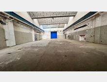 Innenansicht einer großen, leerstehenden Industrielagerhalle mit hohen Decken aus Beton, hellen Backsteinwänden und einem Betonboden mit Kreisspuren. Im Hintergrund befindet sich ein großes blaues Tor sowie teilweise sichtbare Leitungen und technische Installationen an den Wänden.