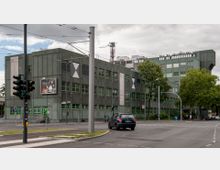 Modernes Bürogebäude mit rechteckiger, dunkelgrüner Fassade und grün gerahmten Fenstern, umgeben von einer Straßenkreuzung mit Ampeln. Im Vordergrund ein parkendes Auto, Plakatwerbung und Bäume entlang der Straße.
