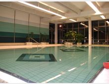 Innenansicht eines Hallenbades mit einem rechteckigen Becken, gefliestem Boden und Handläufen. Im Hintergrund sind bepflanzte Blumenkübel, eine farbige Wandgestaltung in Blautönen sowie große Fensterfronten zu sehen.