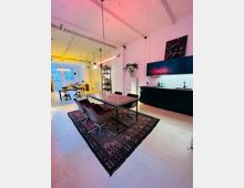 Ein Loft mit offenem Grundriss, hellen Wänden und sichtbaren Deckenbalken. Im Vordergrund ein Esstisch mit Teppich darunter, daneben eine Küche mit dunklen Schränken; im Hintergrund ein Arbeits- oder Wohnbereich mit großen Fenstern und gelber Beleuchtung.