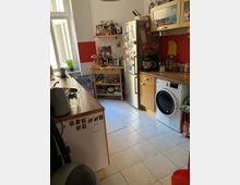 Kleine Küche mit weißen Wänden, roten Akzenten und Fliesenboden. Ausstattung umfasst einen Kühlschrank mit Magneten, eine Waschmaschine, Holzmöbel, offene Regale mit Lebensmitteln und ein großes Fenster, das natürliches Licht hereinlässt.