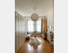 Helles Kinderzimmer mit Holzboden, großen Fenstern mit halbtransparenten Vorhängen und einer zentralen Papierlampe an der Decke. Links befinden sich hohe Schränke, rechts ein Kinderbett, eine Kommode und ein Baldachin, in der Mitte stehen ein kleiner runder Teppich mit einem Kindertisch und Stühlen.