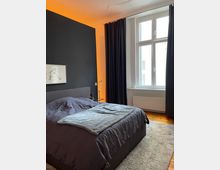 Modernes Schlafzimmer mit dunkler Wand und einem großen Doppelbett mit grauer Bettwäsche. Neben dem Bett steht eine kleine Lampe, der Raum hat ein großes Fenster mit schwarzen Vorhängen und der Boden ist aus Holz mit einem grauen Teppich.