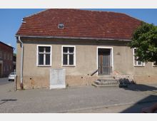 Ein schlichtes, eingeschossiges Haus mit verputzter Fassade und einem roten Satteldach. Das Gebäude hat zwei Fenster mit weißen Rahmen links und eine hölzerne Eingangstür mit kleinen Treppenstufen rechts; daneben steht ein grauer Versorgungskasten.