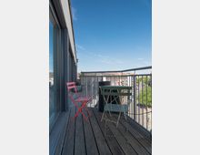 Ein Balkon mit Holzboden, ausgestattet mit einem roten und einem grünen Klappstuhl sowie einem kleinen runden Tisch. Im Hintergrund ein Ausblick auf Dachlandschaften und Bäume, begrenzt durch ein Metallgeländer.