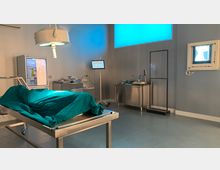 Ein steriler Autopsieraum mit einem mit grünem Tuch bedeckten Untersuchungstisch, einer Deckenleuchte, Edelstahl-Arbeitsflächen, einem Monitor mit der Aufschrift "Team Totenkopf" und einem beleuchteten Röntgenkasten an der Wand. Im Hintergrund sind ein Kühlschrank und medizinische Utensilien sichtbar.