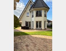 Einfamilienhaus mit hellgelber Fassade und schwarzem Satteldach, ergänzt durch einen runden Erker mit mehreren Fenstern und kleinen Balkonen. Im Vordergrund befinden sich eine gepflasterte Terrasse, ein gepflegter Rasen und ein Gartenstuhl-Set.