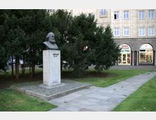 Büste von Karl Marx auf einem weißen Steinsockel in einem kleinen Park mit Rasen und Bäumen im Vordergrund; im Hintergrund befindet sich ein mehrstöckiges Gebäude mit Geschäften im Erdgeschoss.