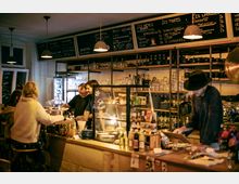Ein gemütliches Café mit einer Theke aus Holz, auf der diverse Getränke und Snacks präsentiert werden. Im Hintergrund sind Regale mit Gläsern, Tassen und Dekoration sowie eine Tafel mit handgeschriebenen Menüangeboten zu sehen, während Gäste und Personal interagieren.