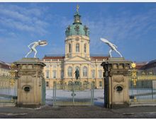Schloss Charlottenburg in Berlin, eine barocke Palastanlage mit einer zentralen Kuppel. Im Vordergrund ein verziertes Tor mit vergoldeten Elementen, flankiert von steinernen Sockeln mit weißen Figuren antiker Krieger.
