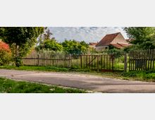 Ein ländlicher Vorgarten mit einem Lattenzaun und einem Metalltor, dahinter dichter Bewuchs aus Bäumen und Sträuchern. Im Hintergrund sind mehrere Häuser mit roten Ziegeldächern zu sehen, daneben ein gepflasterter Gehweg und eine asphaltierte Straße im Vordergrund.