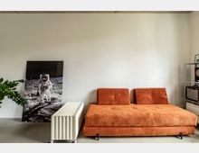 Minimalistisch eingerichteter Raum mit einem orangefarbenen Sofa aus Cordgewebe, einem weißen mobilen Möbelstück und einer auf den Boden gestellten Fotografie eines Astronauten. Im Hintergrund eine weiße Wand, links eine Grünpflanze und rechts ein Regal mit einem Verstärker.