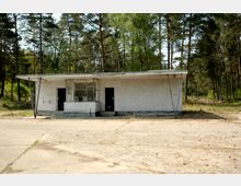 Ein kleines, rechteckiges Gebäude mit flachem Dach und einfacher Betonfassade, das zwei Türen und eine verglaste Fensterfläche aufweist, steht vor einer Umgebung aus Kiefernwald. Der Boden davor besteht aus rissigem Beton, umgeben von Natur.