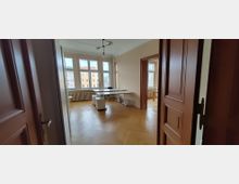 Heller Büroraum mit Fischgrätparkett, hohen Decken und großen Fensterfronten mit Blick auf gegenüberliegende Wohnhäuser. In der Mitte stehen zusammengestellte Schreibtische; dunkle Holztüren und ein Durchgang führen in einen weiteren Raum mit ähnlichen Fenstern und Heizkörpern.