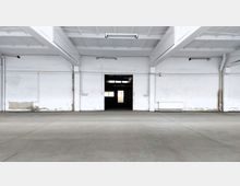 Leerer, weiß gestrichener Industriehallenraum mit einer Betonbodenfläche, einer zentralen offenen Türdurchgangsöffnung und sichtbarer Deckenkonstruktion mit Trägern. Wenige Fenster lassen Licht in den Raum.