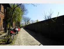 Kopfsteinpflasterstraße mit einer Backsteinmauer auf der rechten Seite und einem Backsteingebäude links. Am Wegrand sind Bäume, ein geparktes Motorrad und Autos zu sehen, im Hintergrund ist ein Tor erkennbar.