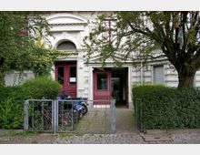 Mehrfamilienhaus mit einer verzierten weiß gestrichenen Fassade und dunkelroten Doppeltüren als Eingänge. Vor dem Gebäude stehen Fahrräder und ein Motorroller hinter einer niedrigen Metallzaunbegrenzung, umgeben von Büschen und einem Baum.