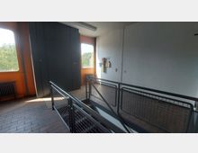 Treppenhaus mit metallenen Handläufen und Gittergeländern, umgeben von weißen und orangefarbenen Wänden. Auf der rechten Seite befinden sich eine Pinnwand, ein Feuerlöscher und ein lichtdurchflutetes Fenster mit Blick ins Grüne.