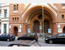 Fassade eines historischen Gebäudes aus rotem Backstein mit großen Rundbögen, verzierten Fenstern und einem zentralen Tor, das von einem schmiedeeisernen Zaun geschützt wird. Mehrere geparkte Autos und ein Fahrrad stehen entlang der Straße, und Plakate hängen an der Fassade und dem Zaun.