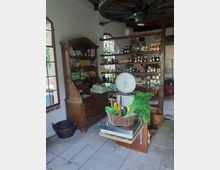 Ein kleiner, rustikaler Laden mit weißen Wänden, Holzbalken und großen Fenstern. Es gibt Regale mit Konserven und Produkten, eine antike Waage mit einem Korb frischen Gemüses sowie eine Holzvitrine mit Lebensmitteln und einer alten Registrierkasse.