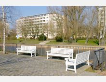Gepflasterte Uferpromenade mit weißen Bänken und einem Geländer, dahinter ein Fluss. Im Hintergrund stehen laublose Bäume und ein mehrstöckiges Wohngebäude.
