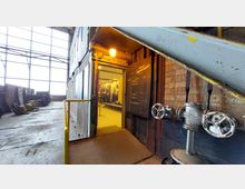 Industriegebäude mit Ziegelwänden und großer Fensterfront. Im Vordergrund ein geöffneter Eingang mit einem beleuchteten Raum dahinter, daneben eine Tafel mit Aufschrift, sowie metallische Ventile und Rohre.