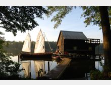 Holzhaus mit Wellblechdach am Ufer eines ruhigen Sees, neben dem zwei Segelboote mit weißen Segeln festgemacht sind. Ein Steg führt vom Ufer zu dem Haus, umgeben von Bäumen und Spiegelungen im Wasser.