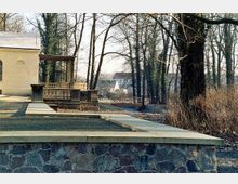 Teilansicht eines historischen Parks mit einer steinernen Balustrade, flankiert von Bäumen mit kahlen Ästen. Im Hintergrund ist ein kleiner Holzsteg über einem Bach sowie Gebäude eines Dorfes zu sehen.