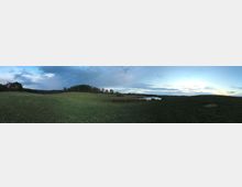 Panorama einer offenen, leicht hügeligen Wiesenlandschaft mit einzelnen Baumgruppen und einem kleinen Teich in der Mitte. Im Hintergrund sind ein dichter Wald und ein leicht bewölkter Himmel bei Sonnenuntergang sichtbar.