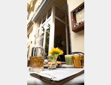 Ein kleiner Tisch in einem Café mit einer Glasflasche Wasser, einer Sonnenblume in einem Glas, einem grünen Getränk und Gebäck auf einem Teller. Im Hintergrund sind eine helle Hausfassade und große Fenster zu sehen.
