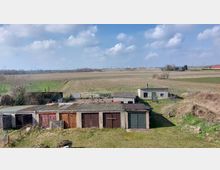 Ländliche Szene mit flachen Garagen und kleinen Nebengebäuden aus Beton im Vordergrund, umgeben von einem weitläufigen, offenen Feld und Wiesen. Im Hintergrund sind Strommasten, Bäume und vereinzelte Gebäude unter einem leicht bewölkten Himmel erkennbar.