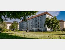 Wohnsiedlung mit mehrstöckigen, hellen Mehrfamilienhäusern und roten Dächern, umgeben von einer Grünfläche mit Bäumen, Wäscheleinen und einem Rasenbereich. Im Vordergrund steht eine Grünfläche mit blühendem Gras und Schatten von Bäumen.