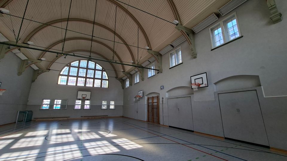 Innenansicht einer großen Turnhalle mit hoher Tonnengewölbe-Decke, Oberlichtfenstern und einem großen Rundbogenfenster an der Stirnseite. An den hellgrauen Wänden hängen mehrere Basketballkörbe; auf dem Hallenboden sind Spielfeldlinien markiert und seitlich stehen Bänke sowie eine Sprossenwand.