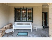 Überdachter Bereich mit einer weißen Holzbank, kleinem weißen Beistelltisch und rundem Metalltisch. Im Hintergrund ein vergittertes Fenster und ein offener Eingang, rechts gestapeltes Holz und ein Glasbodenfenster im Boden.