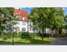 Wohnstraße mit mehrstöckigem Wohnhaus im Hintergrund, das ein hell verputztes Äußeres und ein rotes Ziegeldach aufweist. Vor dem Gebäude stehen Bäume und Sträucher, ein Straßenschild mit den Namen "Alemannenstraße" und "Cimbernstraße" befindet sich im Vordergrund.