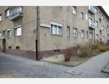 Mehrstöckiges Wohngebäude aus Putz mit schlichten Fassaden und einigen Balkonen, flankiert von einem gepflasterten Gehweg und einem schmalen Grünstreifen mit kahlen Sträuchern. Die Ecke des Gebäudes trägt ein Straßenschild mit der Aufschrift "Germaniagarten 1-2".