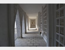 Ein überdachter Gang mit gemauerten Bögen auf der linken Seite und einer Wand mit Nischen, die Namen und Daten zeigen, auf der rechten Seite. Im Hintergrund steht eine weiße Bank, und einzelne Blumen befinden sich in den Nischen.